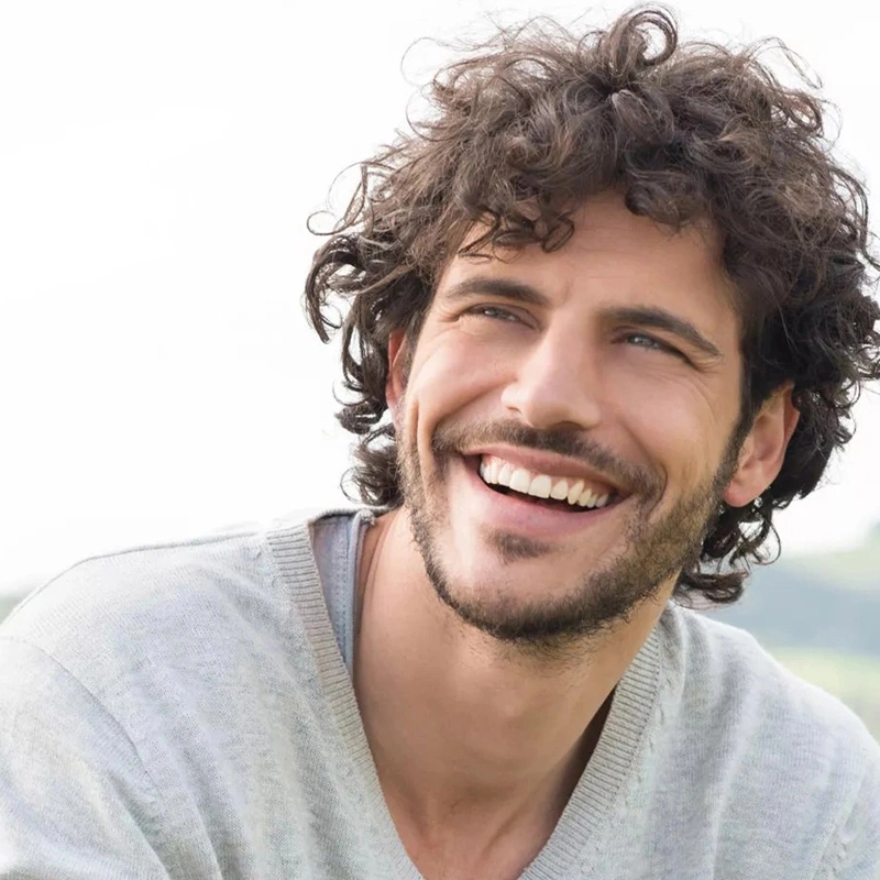 Smiling man with curly brown hair wearing light gray shirt against blurred outdoor background.
