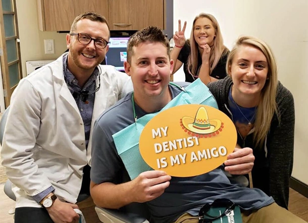 Dental team members hold "My Dentist is My Amigo" sign in office setting.
