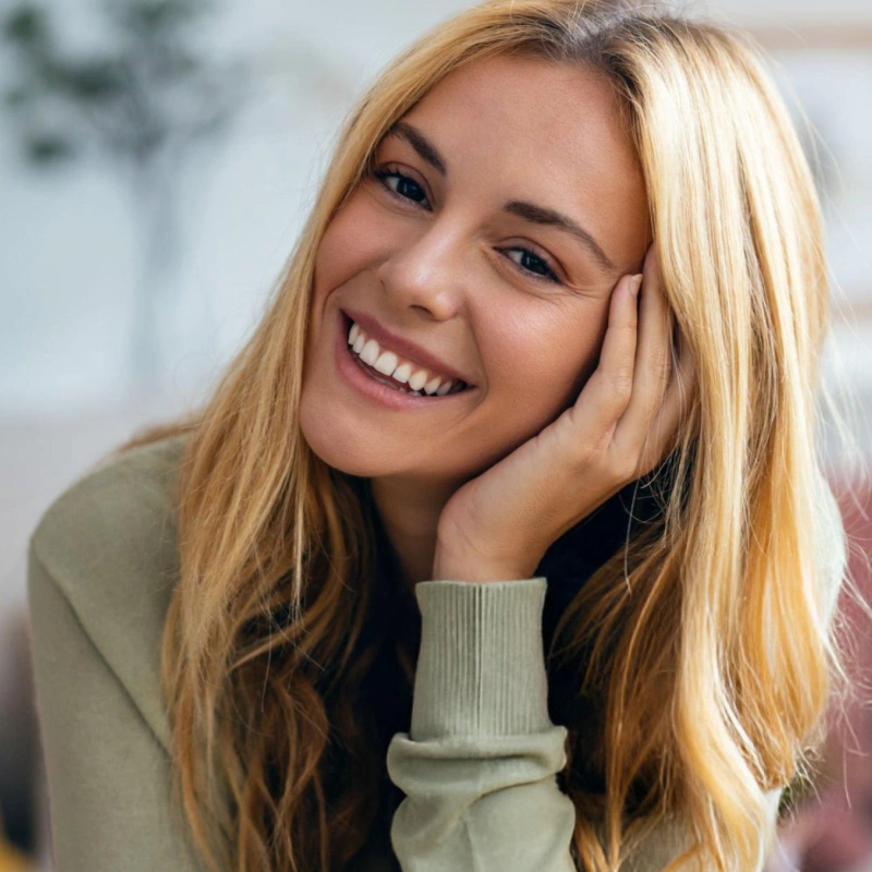 Young woman smiling warmly at camera, hand near face, wearing sage green sweater indoors.